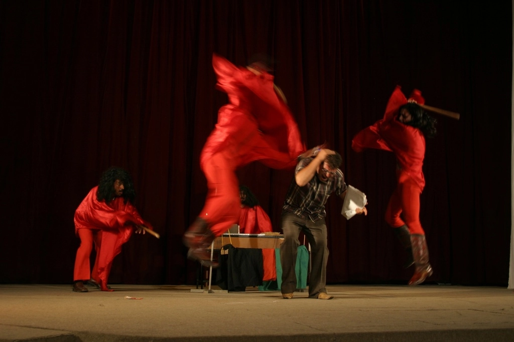 Photo d'archive d'Afghanistan. Compagnie Hasards d'Hasards. Photo de représentation de spectacle. Sur scène, trois comédiens habillés de rouge frappe un autre comédiens avec des bâtons.
