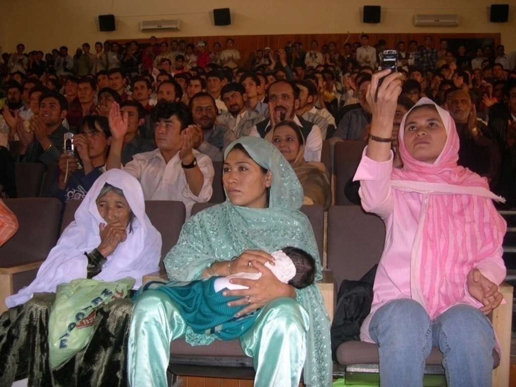 Photo d'archive d'Afghanistan. Compagnie Hasards d'Hasards. Photo du public. Trois femmes au premier rang. L'une d'elle porte un bébé dans ses bras. Une autre essais de capter du réseaux avec sont téléphone portable.